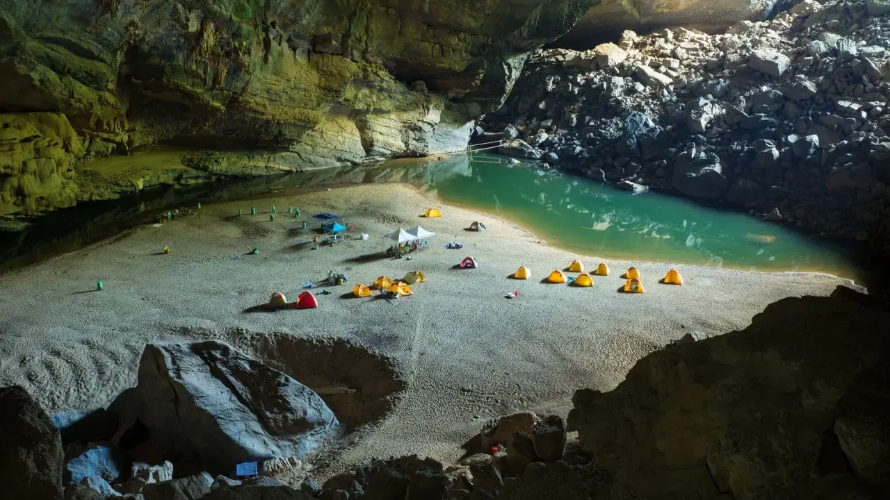 Obóz w jaskini Son Doong: namioty rozbite na piaszczystej plaży nad turkusową wodą, otoczone skalnymi ścianami.