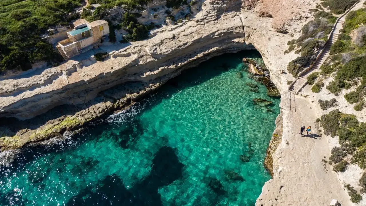 Malta, gdzie na malta? Widok z lotu ptaka na turkusową zatokę, klify i mały domek. Dwie osoby spacerują ścieżką.