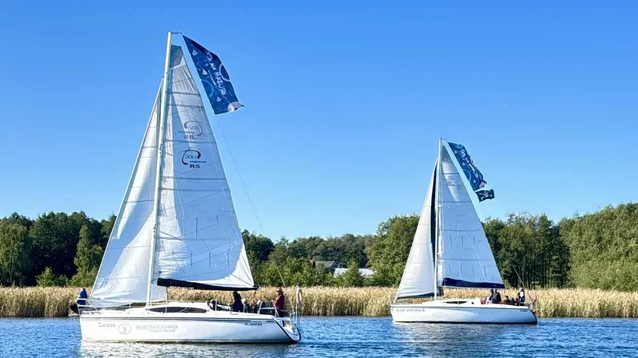 Dwa żaglowce na jeziorze, symbolizujące to, z czego słyną Mazury – żeglarstwo i piękne krajobrazy.
