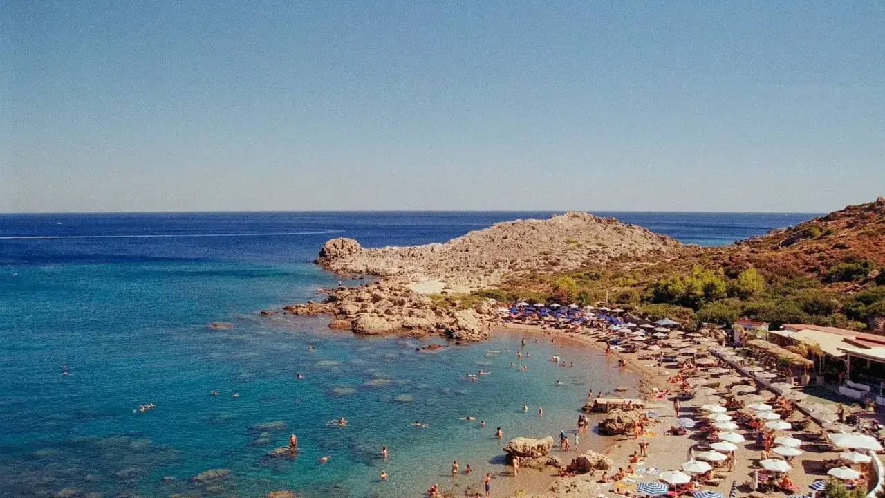 Rodos: turkusowe morze Egejskie, kamienista plaża z leżakami i parasolami, ludzie kąpiący się w wodzie.