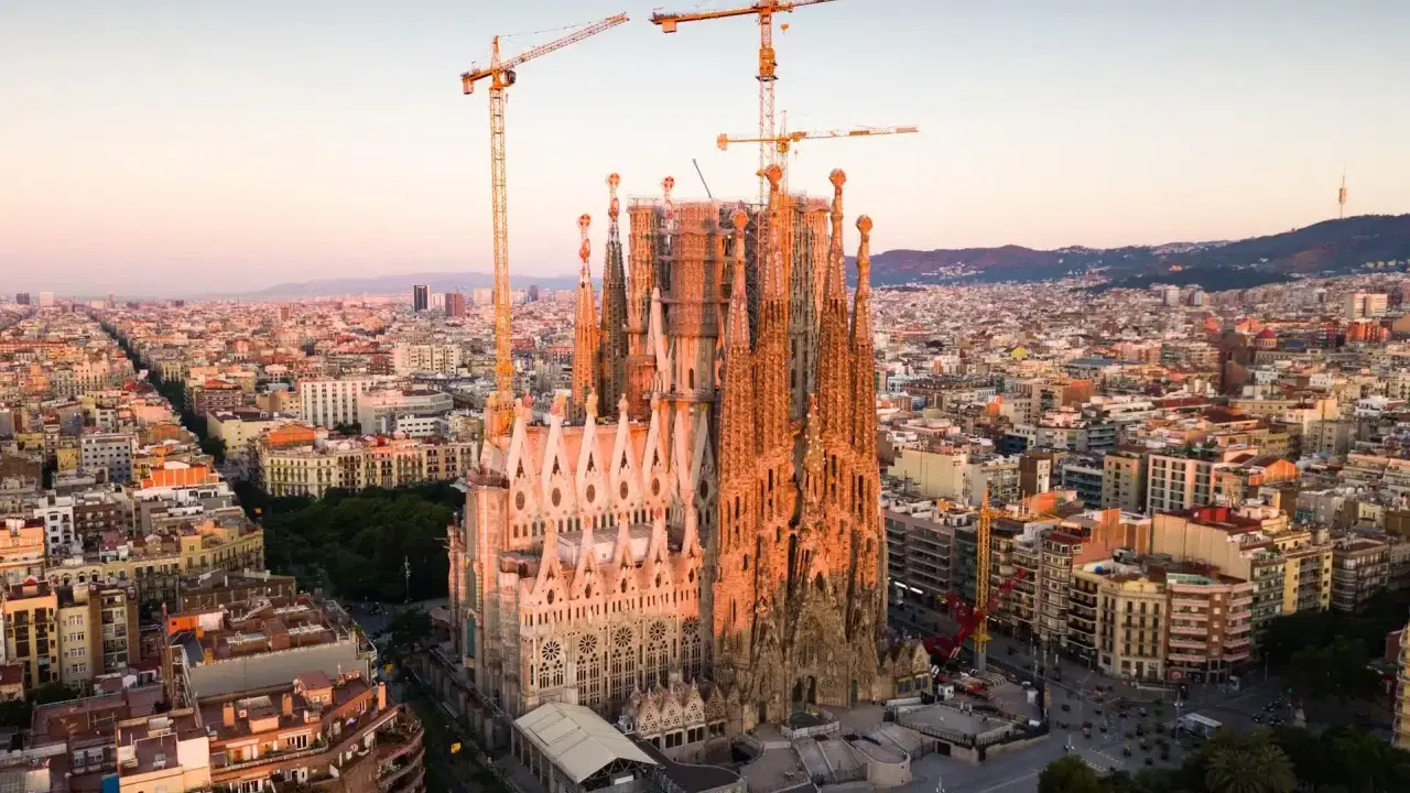 Widok z lotu ptaka na Sagrada Familia w Barcelonie, wciąż w budowie, z dźwigami górującymi nad miastem. Słońce oświetla fasadę, tworząc ciepłą poświatę, jakby przygotowując się do wieczornych mszy.