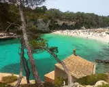 Majorka: turkusowa woda, piaszczysta plaża pełna ludzi i parasoli, skaliste klify, sosny i mały domek z czerwoną dachówką.