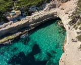 Malta, gdzie na malta? Widok z lotu ptaka na turkusową zatokę, klify i mały domek. Dwie osoby spacerują ścieżką.