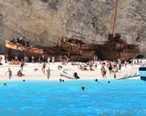 Słynna zatoka wraku zamknięta dla turystów, ale plaża tętni życiem. Ludzie kąpią się w turkusowej wodzie i odpoczywają na piasku obok zardzewiałego statku.