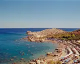 Rodos: turkusowe morze Egejskie, kamienista plaża z leżakami i parasolami, ludzie kąpiący się w wodzie.
