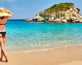 Kobieta w kapeluszu i bikini podziwia turkusowe morze i skalistą wysepkę. Idealne plaże w Grecji.