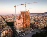Widok z lotu ptaka na Sagrada Familia w Barcelonie, wciąż w budowie, z dźwigami górującymi nad miastem. Słońce oświetla fasadę, tworząc ciepłą poświatę, jakby przygotowując się do wieczornych mszy.