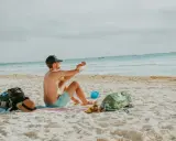 Mężczyzna robi zdjęcie telefonem na plaży, marząc o najkrótszych lotach z Polski na wakacje. Obok leżą torba, kapelusz i sandały.