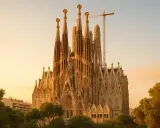 Sagrada Familia w Barcelonie, wciąż w budowie, góruje nad drzewami w złotym świetle zachodzącego słońca. Dźwig budowlany przypomina o trwającym dziele.