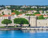 Kamienne mury i wieże otaczają urokliwe, stare miasto Rodos. W tle zielone wzgórza i białe budynki.