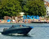Motorówka sunie po wodzie, w tle plaża pełna ludzi i wielki napis "GIŻYCKO". Idealny dzień na Mazurach.