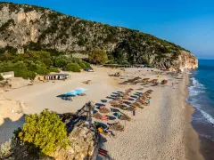 Plaża z leżakami i parasolami pod skalistym klifem, nad turkusowym morzem.