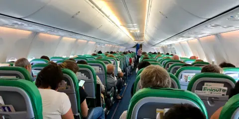 Wnętrze samolotu Transavia, pasażerowie siedzą w rzędach, przygotowani do lotu. Bagaż podręczny schowany w schowkach nad głowami.