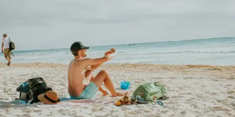 Mężczyzna robi zdjęcie telefonem na plaży, marząc o najkrótszych lotach z Polski na wakacje. Obok leżą torba, kapelusz i sandały.