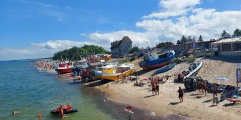 Słoneczny dzień na plaży, kolorowe łodzie na piasku, tłumy ludzi szukają idealnych miejsc na wakacje.
