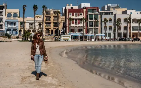 Kobieta spaceruje po piaszczystej plaży w pretty bay Malta, w tle kolorowe budynki i palmy.