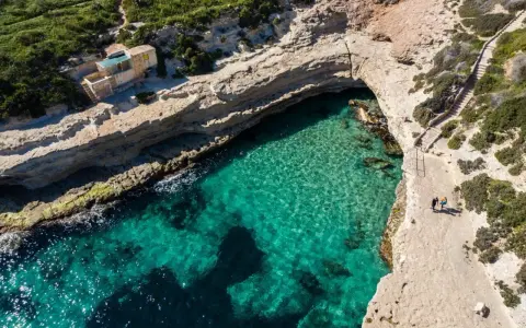Malta, gdzie na malta? Widok z lotu ptaka na turkusową zatokę, klify i mały domek. Dwie osoby spacerują ścieżką.