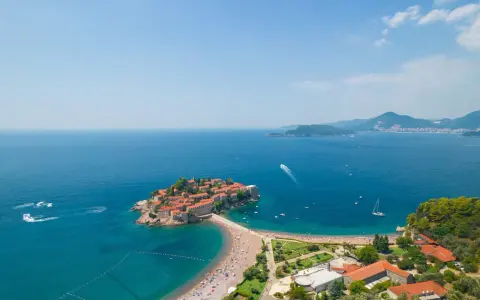Idealna czarnogóra pogoda wrzesień: słoneczna wyspa Sveti Stefan, turkusowe morze, plaża pełna ludzi i łodzie na wodzie.