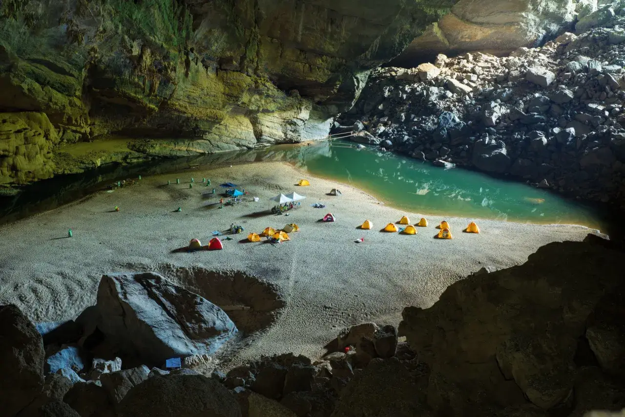 Obóz w jaskini Son Doong: namioty rozbite na piaszczystej plaży nad turkusową wodą, otoczone skalnymi ścianami.
