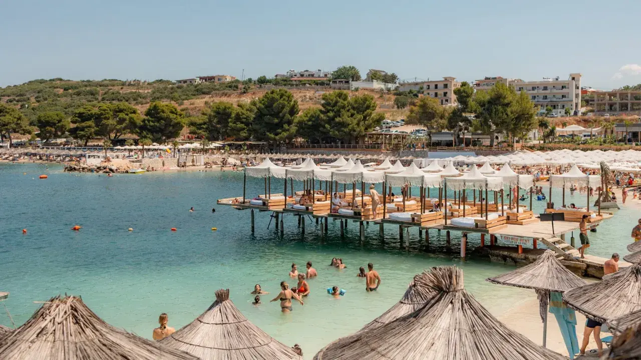 Słoneczny dzień w Ksamil Albania. Ludzie relaksują się na plaży i w wodzie, a nad zatoką unoszą się białe parasole.