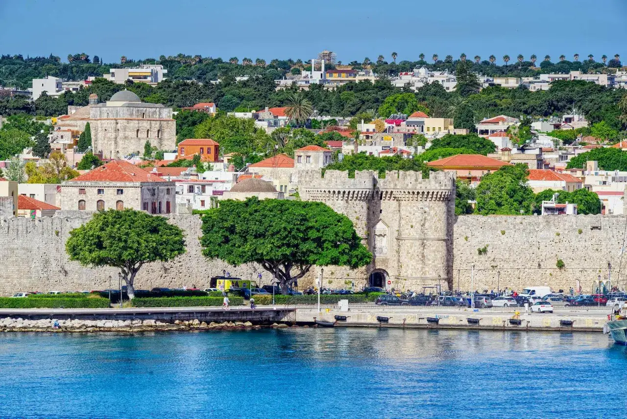 Kamienne mury i wieże, zielone drzewa i niebieska woda tworzą malowniczy widok na stare miasto Rodos.