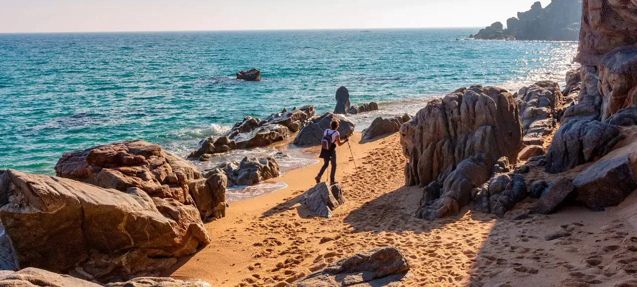 Kobieta z kijkami trekkingowymi idzie piaszczystą plażą wzdłuż skalistego wybrzeża. To piękny fragment szlaku camino de ronda.