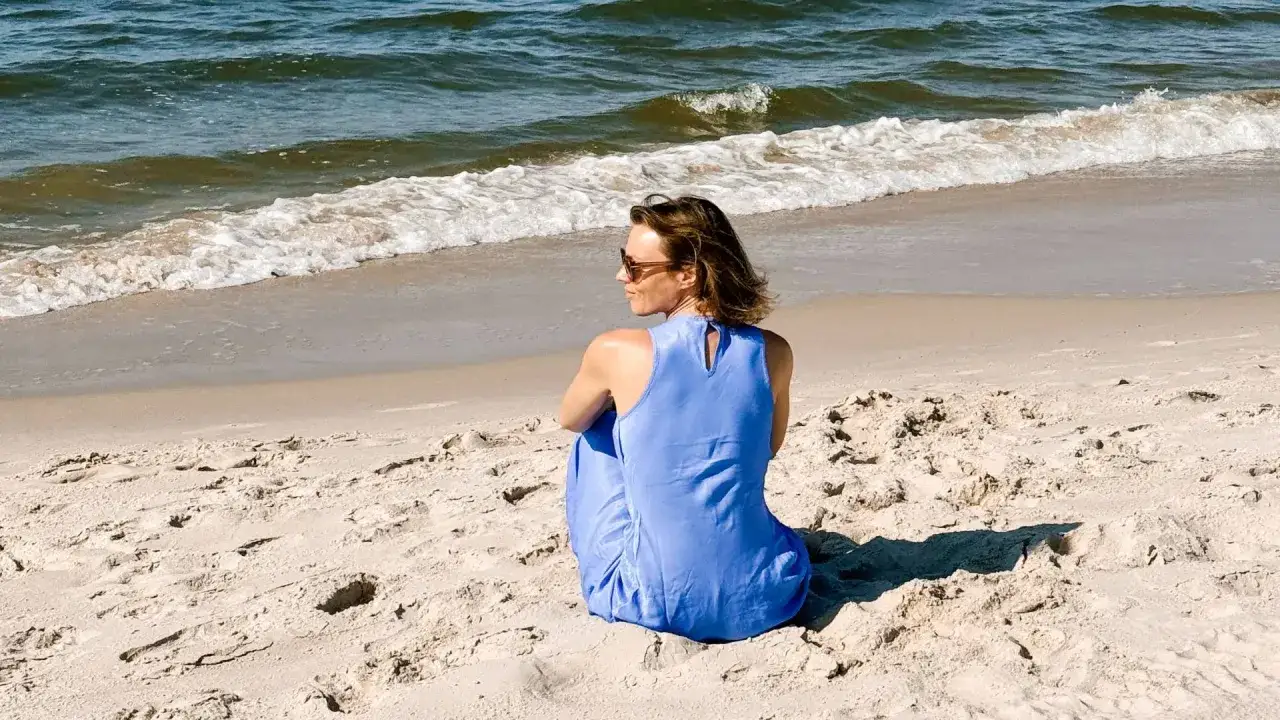 Kobieta w niebieskiej sukience siedzi na piaszczystej plaży, patrząc na fale. Idealne miejsce, gdzie warto polecieć na wakacje.
