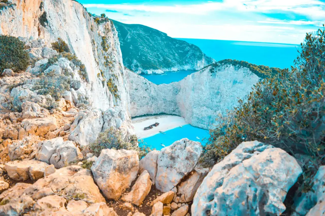 Widok na zatokę wraku na Zakynthos. Białe klify otaczają piaszczystą plażę i turkusową wodę, na której widać wrak statku i łodzie.