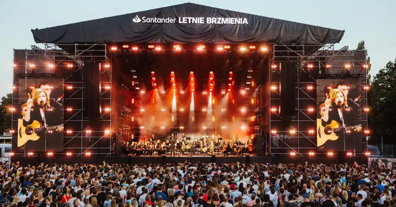 Koncert na scenie Letnie Brzmienia, jednym z corocznych festiwali w Polsce. Tłum ludzi, artystka z gitarą na telebimie.