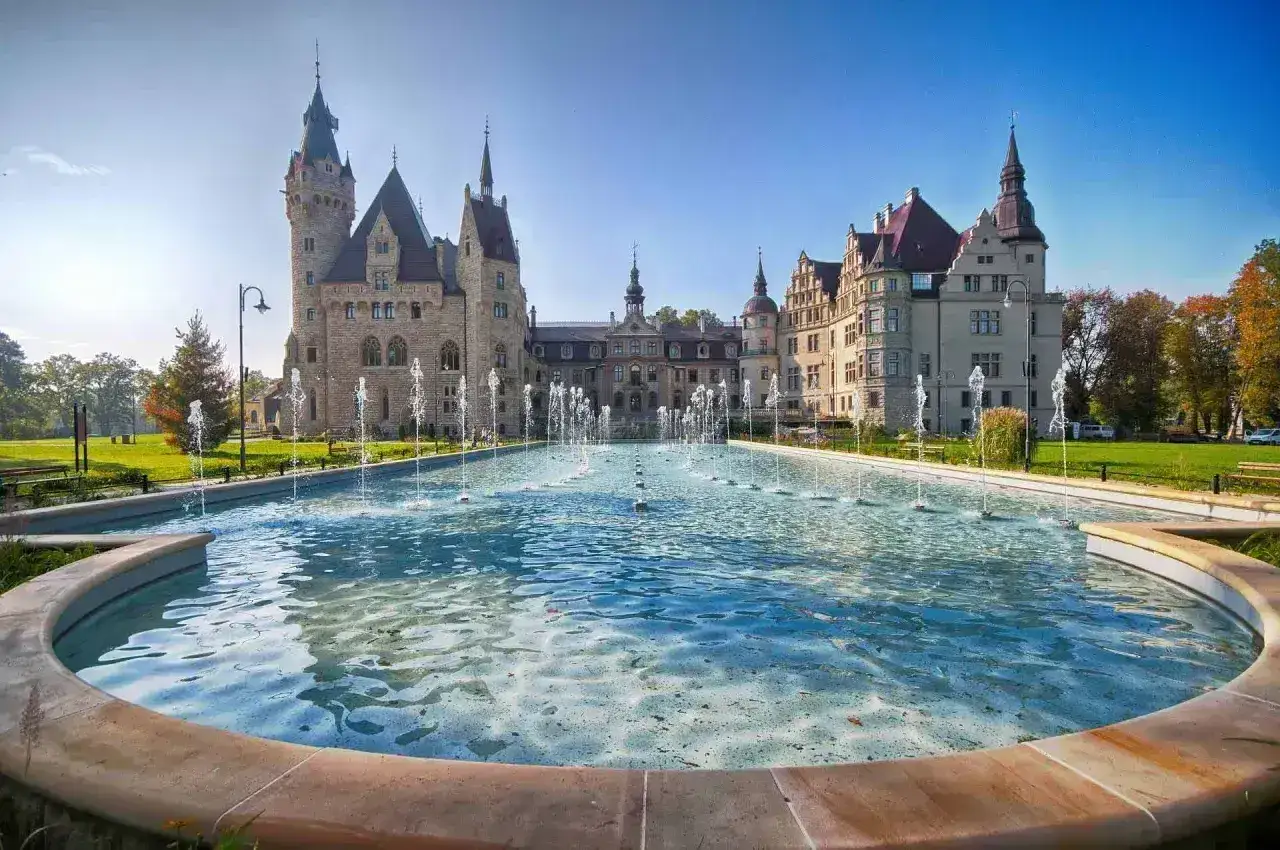 Majestatyczny pałac w Mosznej z licznymi wieżami i bogatą architekturą, otoczony zielenią i ozdobiony kaskadą fontann.