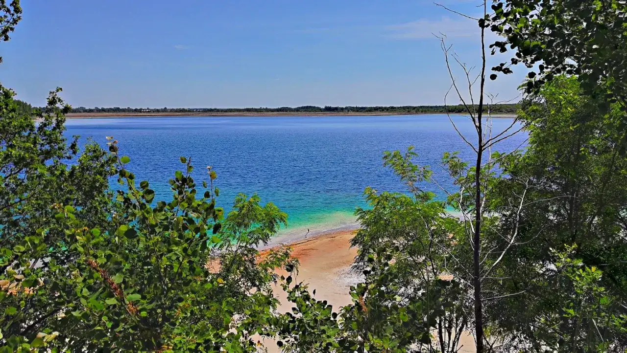 Piękny widok na turkusowe jezioro z piaszczystą plażą, otoczone zielenią. Idealne miejsce na spacer, jak z bajki, gdzieś w pobliżu osadnika gajówka.
