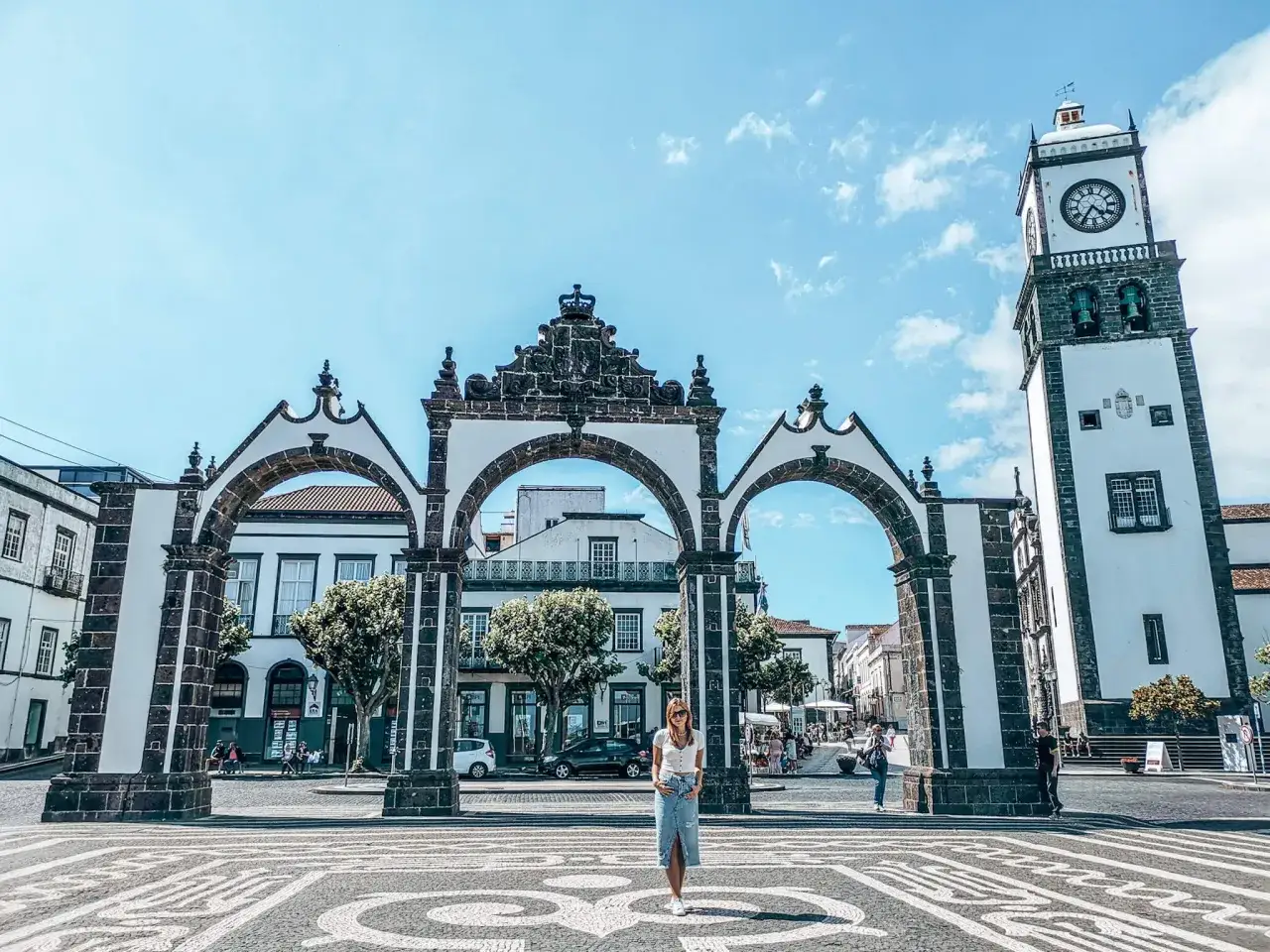 Kobieta na tle bramy miejskiej w Ponta Delgada, Azory. W tle wieża zegarowa.