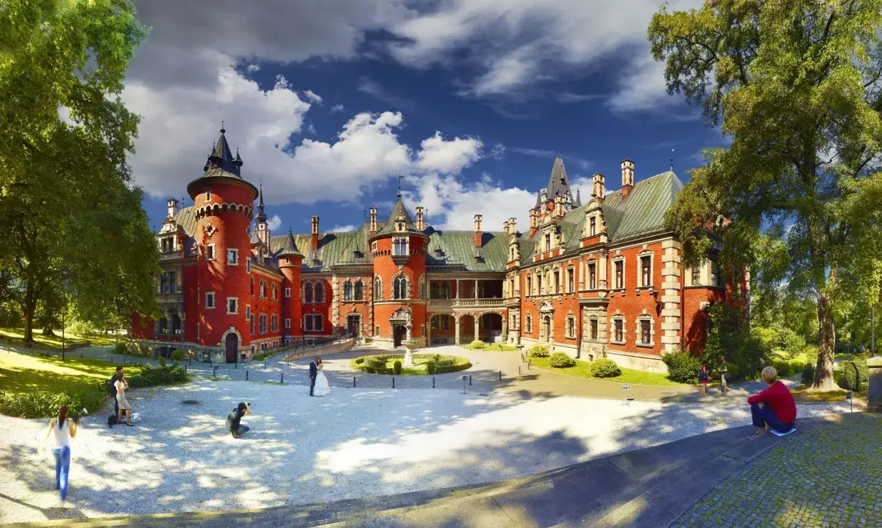 Czerwony zamek z wieżyczkami, idealne miejsce, by zobaczyć, co warto zobaczyć na Śląsku. Wokół zielony park, a na dziedzińcu sesja ślubna.