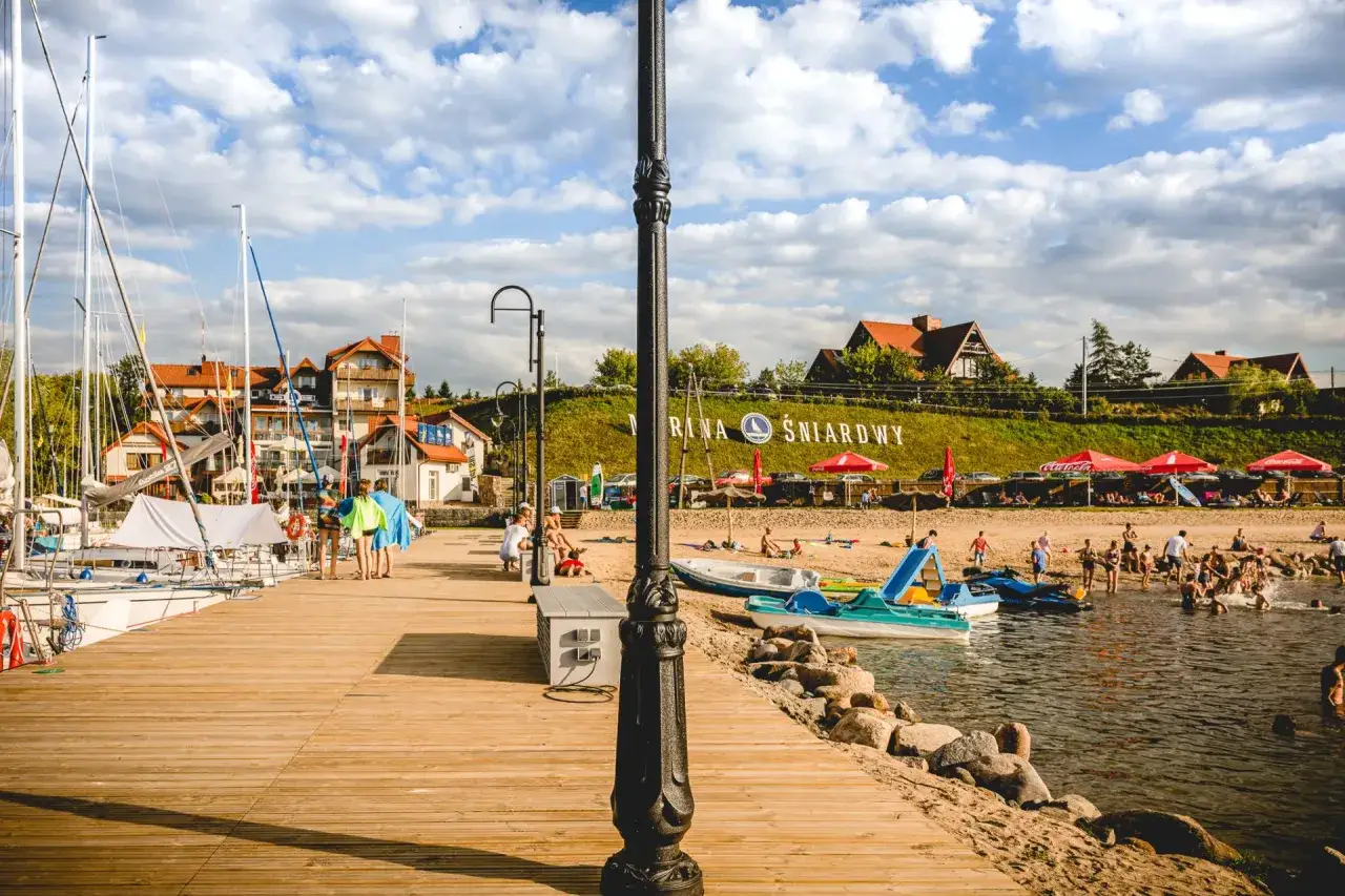 Letnie popołudnie nad jeziorem. Drewniany pomost, żaglówki, plaża z parasolami i ludzie cieszący się wodą. To są fajne miejsca nad jeziorem.
