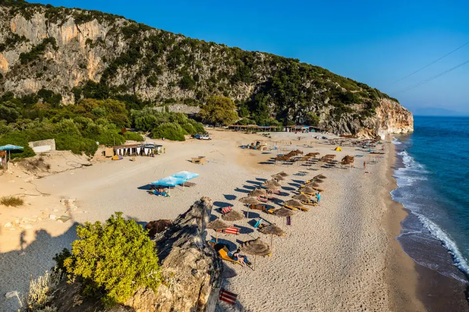 Plaża z leżakami i parasolami pod skalistym klifem, nad turkusowym morzem.